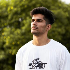Young man in a white 'Street Jutsu' shirt looking upwards outdoors, with blurred green background.